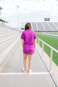 Woman with dark hair wearing the Daisy Top and Jane Skirt set in a back profile position at a football field. The focal point of this image is the backside of the set showing intricate pattern on fabric as well as stripes on cuff of shirt. The model paired this with neutral sneakers.