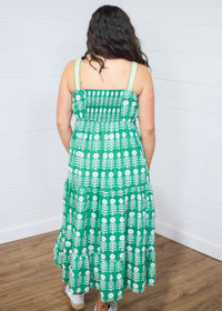 Woman with dark hair standing in a back profile position wearing the Demi Dress against a white background. The focal point of this image is graphics on back, yellow chevron straps as well as the scrunched stretched area on back. The model paired this dress with white moccasins. 