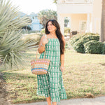 Woman with dark hair in a front profile position wearing the Demi Dress in a lifestyle scene. The focal point of this image is the dress in its entirety. The model paired this dress with white strappy heels and a woven hand-bag. 