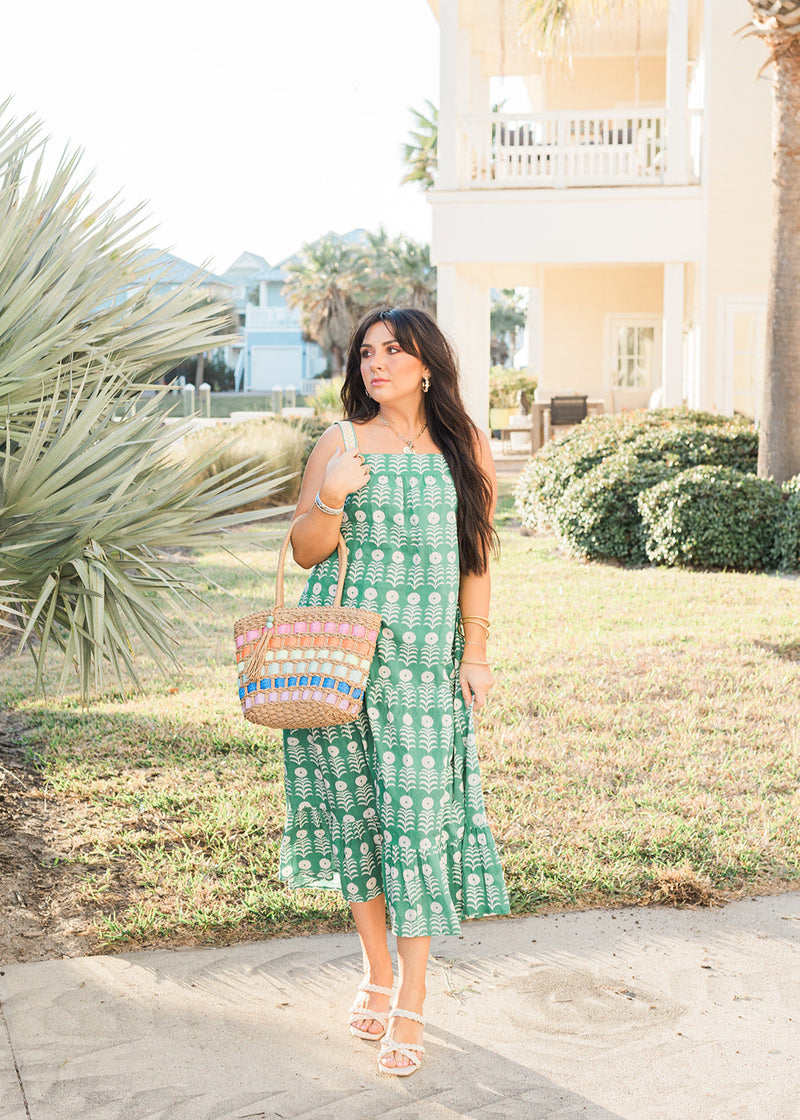 Woman with dark hair in a front profile position wearing the Demi Dress in a lifestyle scene. The focal point of this image is the dress in its entirety. The model paired this dress with white strappy heels and a woven hand-bag. 