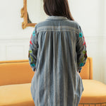 Woman with dark hair standing in a back profile position wearing the Diane Top in a lifestyle scene. The focal point of this image is the denim fabric and sneak peek of floral embroidery on sleeve. The model paired this top with mid-wash denim jeans. 