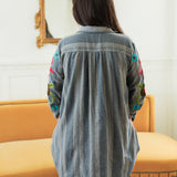 Woman with dark hair standing in a back profile position wearing the Diane Top in a lifestyle scene. The focal point of this image is the denim fabric and sneak peek of floral embroidery on sleeve. The model paired this top with mid-wash denim jeans. 
