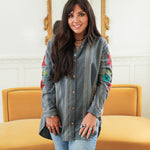 Woman with dark hair standing in a front profile position wearing the Diane Top in a lifestyle scene. The focal point of this image is the denim fabric, floral embroidery, and buttons down front of the top. The model paired this top with mid-wash denim jeans. 