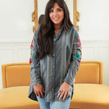Woman with dark hair standing in a front profile position wearing the Diane Top in a lifestyle scene. The focal point of this image is the denim fabric, floral embroidery, and buttons down front of the top. The model paired this top with mid-wash denim jeans. 