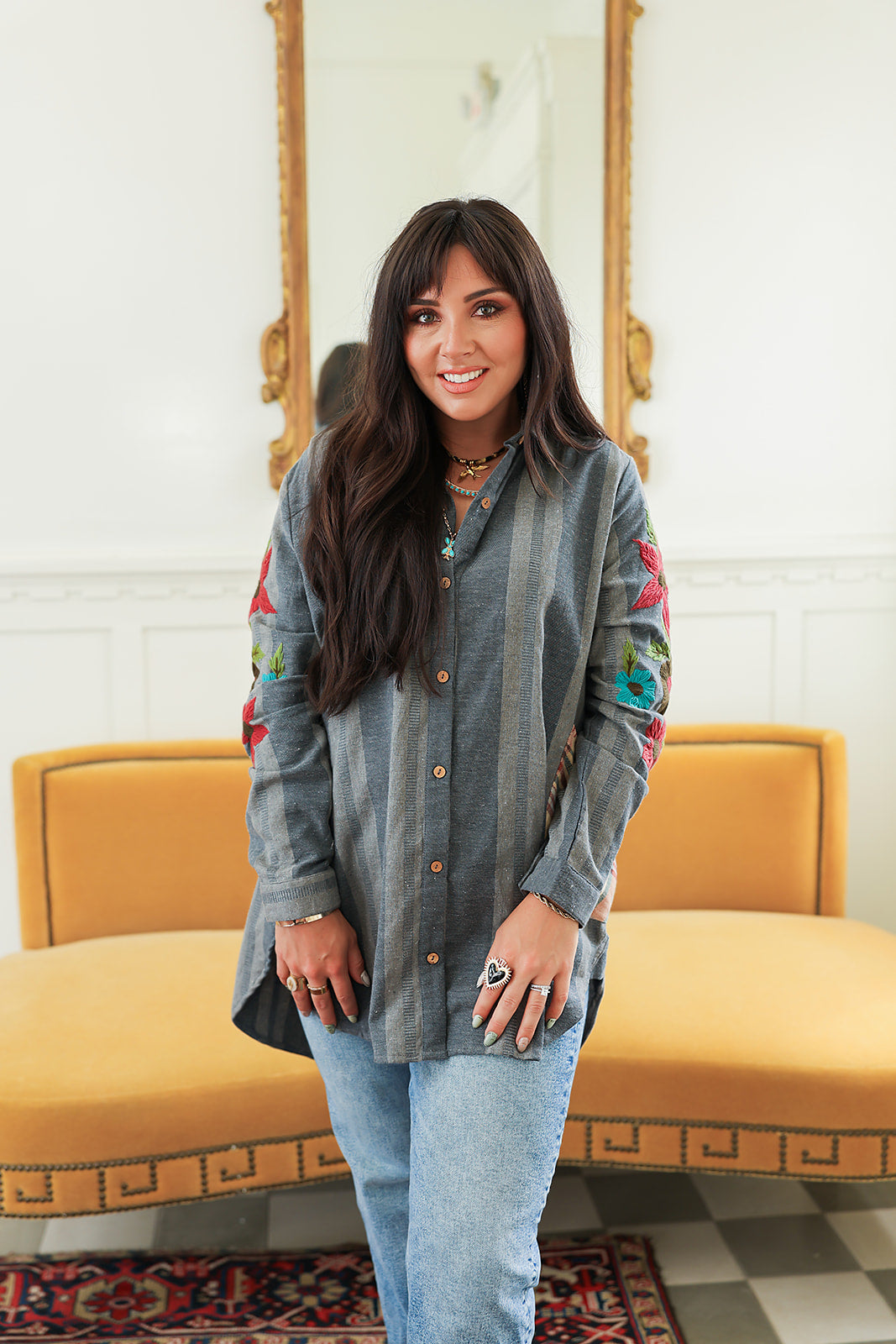 Woman with dark hair standing in a front profile position wearing the Diane Top in a lifestyle scene. The focal point of this image is the denim fabric, floral embroidery, and buttons down front of the top. The model paired this top with mid-wash denim jeans. 