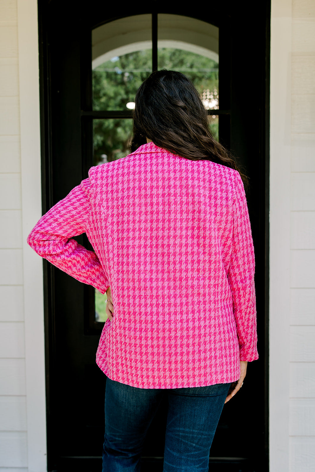 Woman with dark hair wearing a pink tweed blazer with gold buttons. Model paired blazer with dark-wash denim jeans. 
