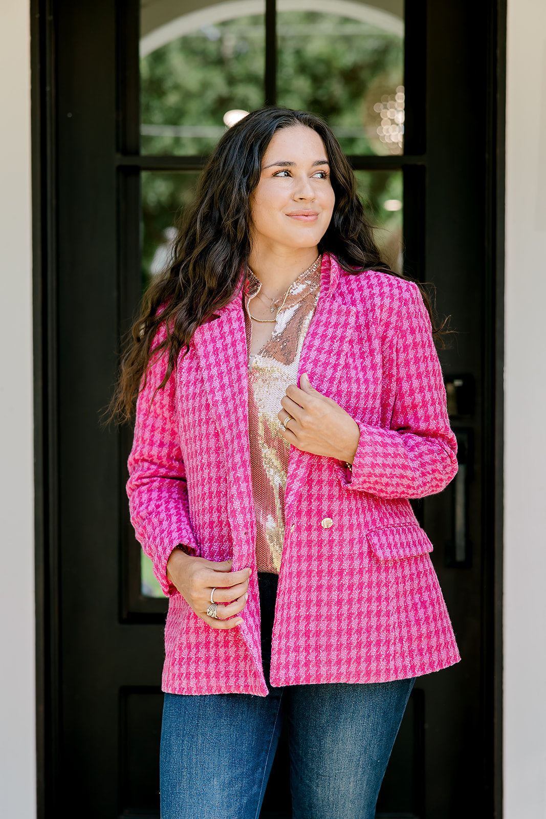 Woman with dark hair wearing a pink tweed blazer with gold buttons. Model paired blazer with dark-wash denim jeans. 