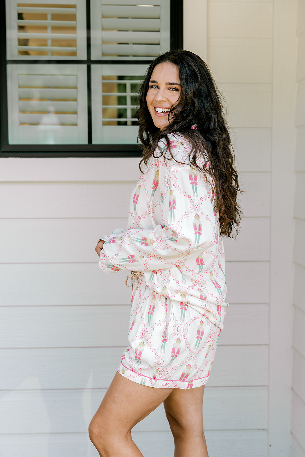 Woman with dark hair wearing a long-sleeve top and shorts pajama set featuring nutcracker graphics. 