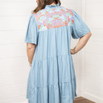 Woman with dark hair standing in a back profile position wearing the Evie Dress against a white background. The focal point of this image is the orange, green, and pink embroidery with pleating and ruffles. 