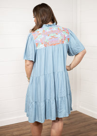 Woman with dark hair standing in a back profile position wearing the Evie Dress against a white background. The focal point of this image is the orange, green, and pink embroidery with pleating and ruffles. 