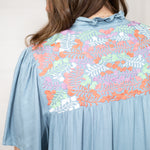 Woman with dark hair standing in a back profile position wearing the Evie Dress  against a white background. The focal point of this image is floral pink, orange, white, and green embroidery. 