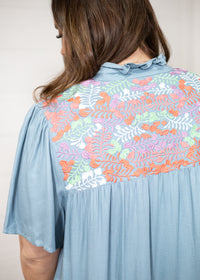 Woman with dark hair standing in a back profile position wearing the Evie Dress  against a white background. The focal point of this image is floral pink, orange, white, and green embroidery. 