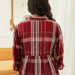 Woman with dark hair standing in a back profile position wearing the Faye Top in a lifestyle scene. The focal point of the image is the maroon plaid graphics on fabric as well as the flowy bottom-half of top. The model paired this top with light-wash denim jeans. 