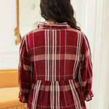 Woman with dark hair standing in a back profile position wearing the Faye Top in a lifestyle scene. The focal point of the image is the maroon plaid graphics on fabric as well as the flowy bottom-half of top. The model paired this top with light-wash denim jeans. 