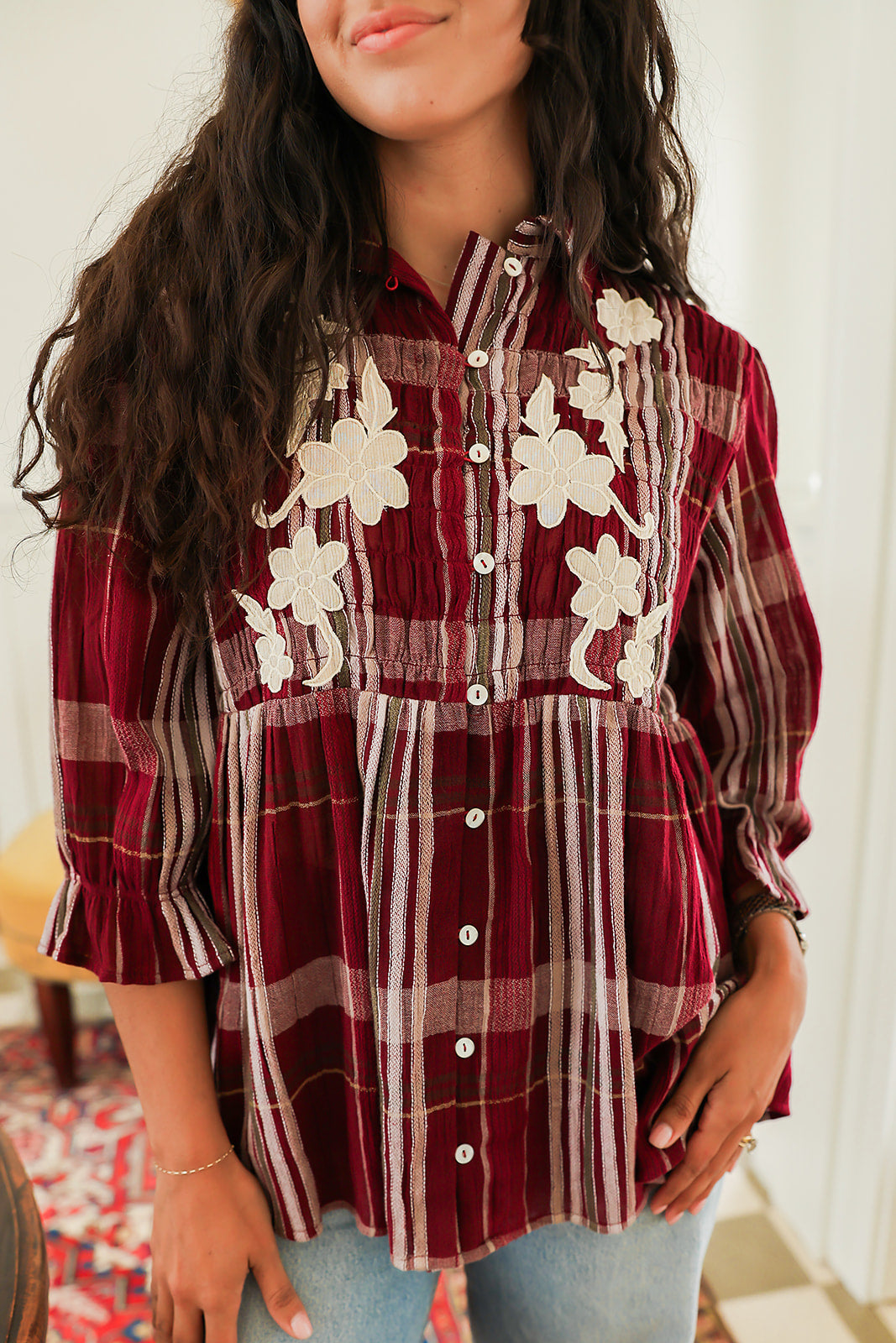 Woman with dark hair standing in a front profile position wearing the Faye Top against a white background. The focal point of this image is the pleating on front of top as well as the buttons and white floral embroidery on chest. The model paired this top with light-wash denim jeans. 