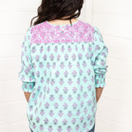 Woman with dark hair standing in a back profile position wearing the Grace Top against a white background. The focal point of this image is the purple and blue floral pattern as well as the purple embroidery across shoulders. The model paired this top with dark-wash denim jeans. 