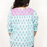 Woman with dark hair standing in a back profile position wearing the Grace Top against a white background. The focal point of this image is the purple and blue floral pattern as well as the purple embroidery across shoulders. The model paired this top with dark-wash denim jeans. 
