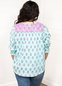 Woman with dark hair standing in a back profile position wearing the Grace Top against a white background. The focal point of this image is the purple and blue floral pattern as well as the purple embroidery across shoulders. The model paired this top with dark-wash denim jeans. 