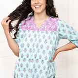 Woman with dark hair standing in a front profile position wearing the Grace Top against a white background. The focal point of this image is the purple embroidery, floral graphics, and v-neckline. The model paired this top with dark-wash denim jeans. 