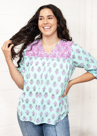 Woman with dark hair standing in a front profile position wearing the Grace Top against a white background. The focal point of this image is the purple embroidery, floral graphics, and v-neckline. The model paired this top with dark-wash denim jeans. 
