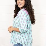Woman with dark hair standing in a side profile position wearing the Grace Top against a white background. The focal point of this image is the purple and mint floral patter with cuffed quarter sleeves. The model paired this top with dark-wash denim jeans. 