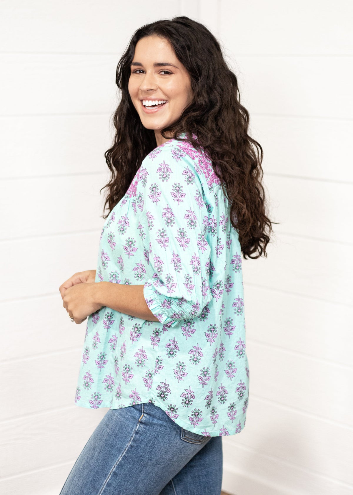 Woman with dark hair standing in a side profile position wearing the Grace Top against a white background. The focal point of this image is the purple and mint floral patter with cuffed quarter sleeves. The model paired this top with dark-wash denim jeans. 