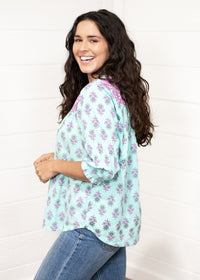 Woman with dark hair standing in a side profile position wearing the Grace Top against a white background. The focal point of this image is the purple and mint floral patter with cuffed quarter sleeves. The model paired this top with dark-wash denim jeans. 