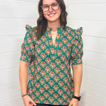 Model with brown hair is standing in a front profile position wearing the Haley Top against a white background. The focal point of this image is the intricate graphics on the top as well as the v-neckline embellished with embroidery. The model paired this top with black pants. 