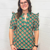 Model with brown hair is standing in a front profile position wearing the Haley Top against a white background. The focal point of this image is the intricate graphics on the top as well as the v-neckline embellished with embroidery. The model paired this top with black pants. 