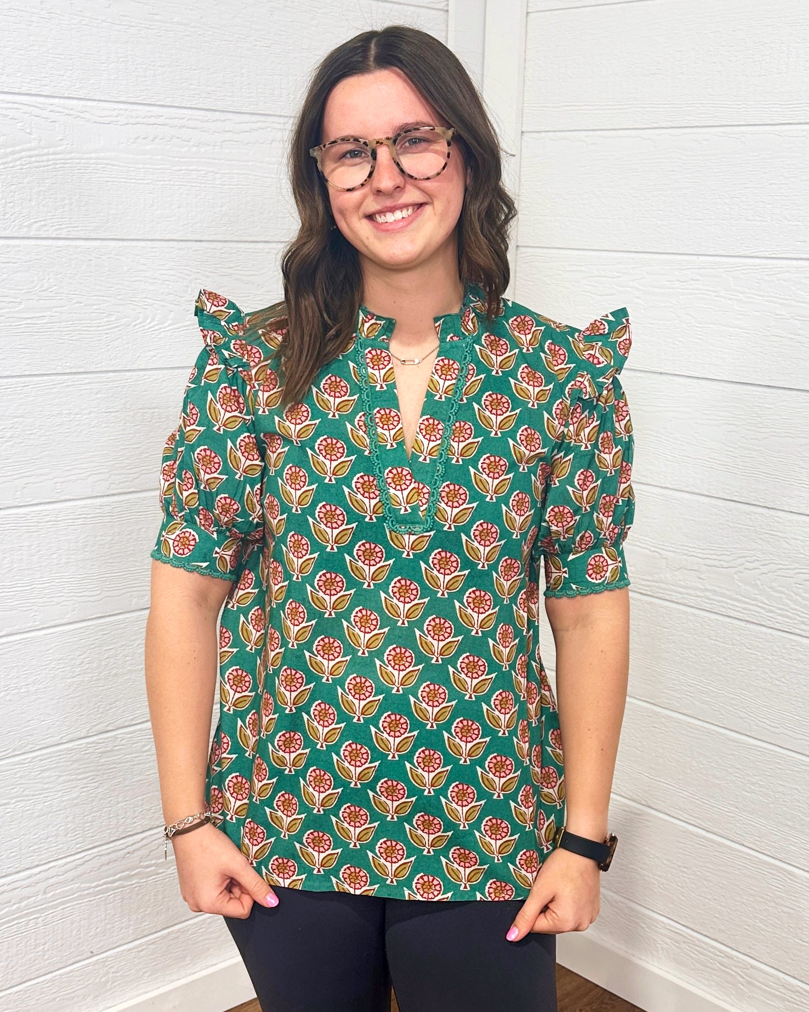 Model with brown hair is standing in a front profile position wearing the Haley Top against a white background. The focal point of this image is the intricate graphics on the top as well as the v-neckline embellished with embroidery. The model paired this top with black pants. 