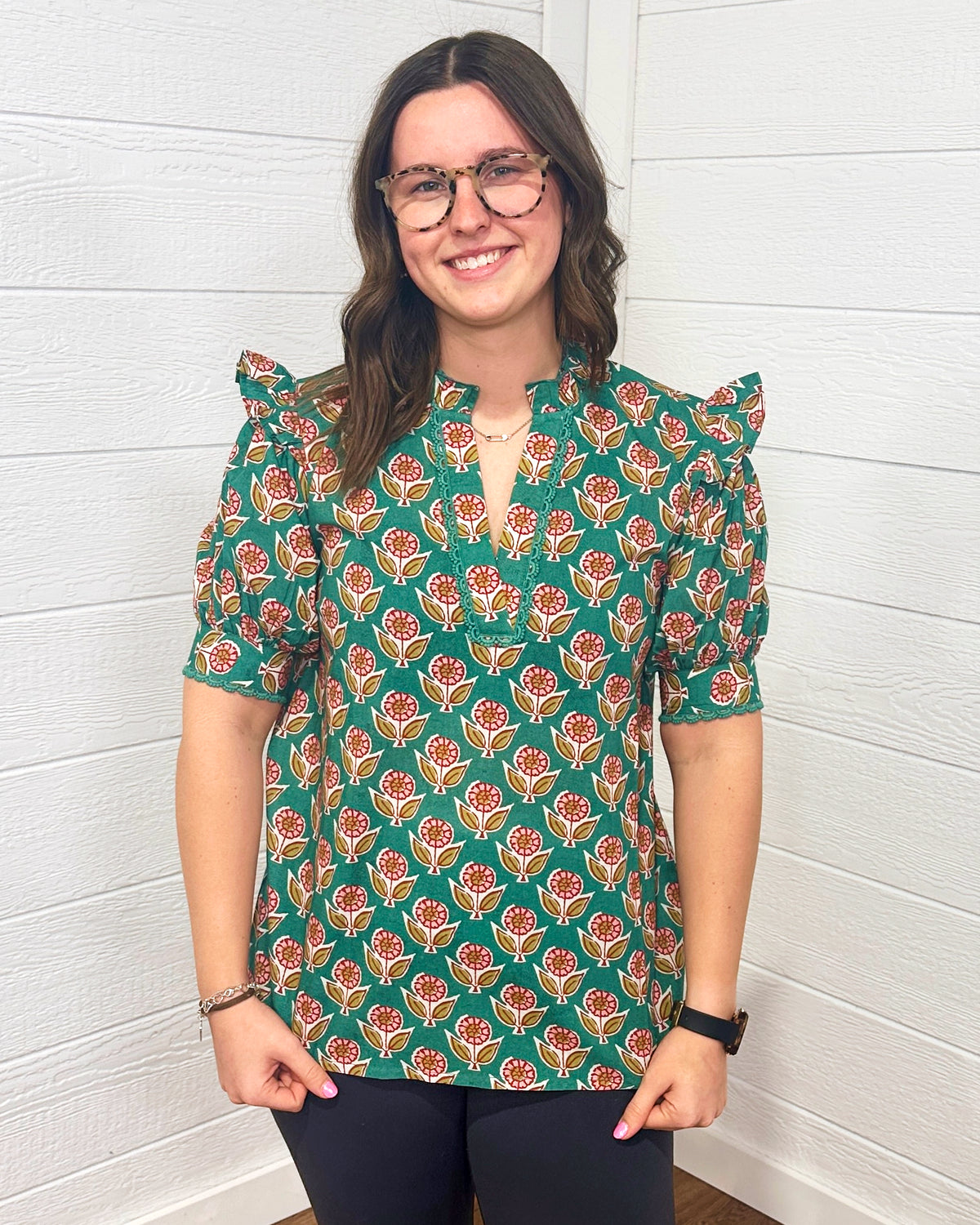 Model with brown hair is standing in a front profile position wearing the Haley Top against a white background. The focal point of this image is the intricate graphics on the top as well as the v-neckline embellished with embroidery. The model paired this top with black pants. 