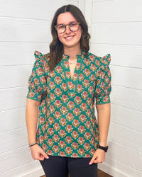 Model with brown hair is standing in a front profile position wearing the Haley Top against a white background. The focal point of this image is the intricate graphics on the top as well as the v-neckline embellished with embroidery. The model paired this top with black pants. 
