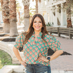 Model with dark hair is standing in a front profile position in a lifestyle scene wearing the Haley Top. The focal point of this image is to see the top in its entirety, that includes all of the fun graphics. The model paired this top with dark-wash denim shorts.
