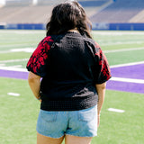 Woman with dark hair wearing a short-sleeve black top with red floral embroidery on sleeves. The model paired this top with light-wash denim shorts. 