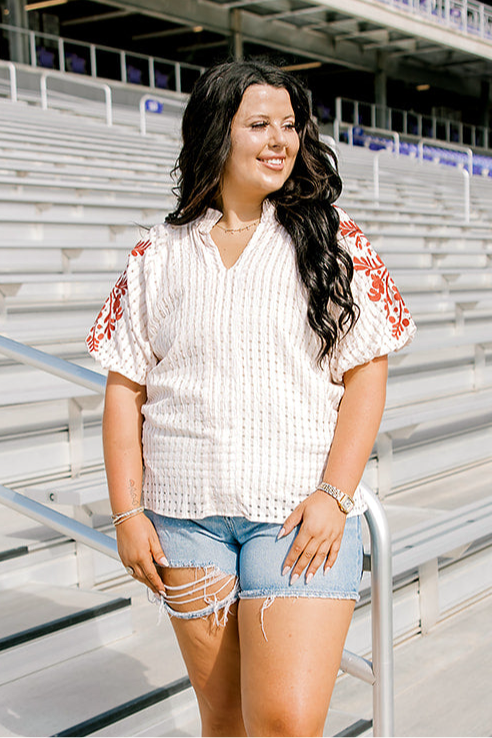 Woman with dark hair wearing a cream short-sleeve top with burnt orange embroidery, and light-wash denim shorts.