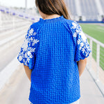 Woman with dark hair standing in a back profile position wearing the Havanah Top in a football field setting. The focal point of this image is the fabric material and white embroidery on the sleeves with slight emphasis on the roused neckline. The model paired these with light-wash denim shorts.
