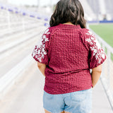 Woman with dark hair standing in a back profile position wearing the Havanah Top at a football field. The focal point of this image is the fabric pattern as well as white floral embroidery down the sleeves. The model paired this top with light-wash denim shorts. 