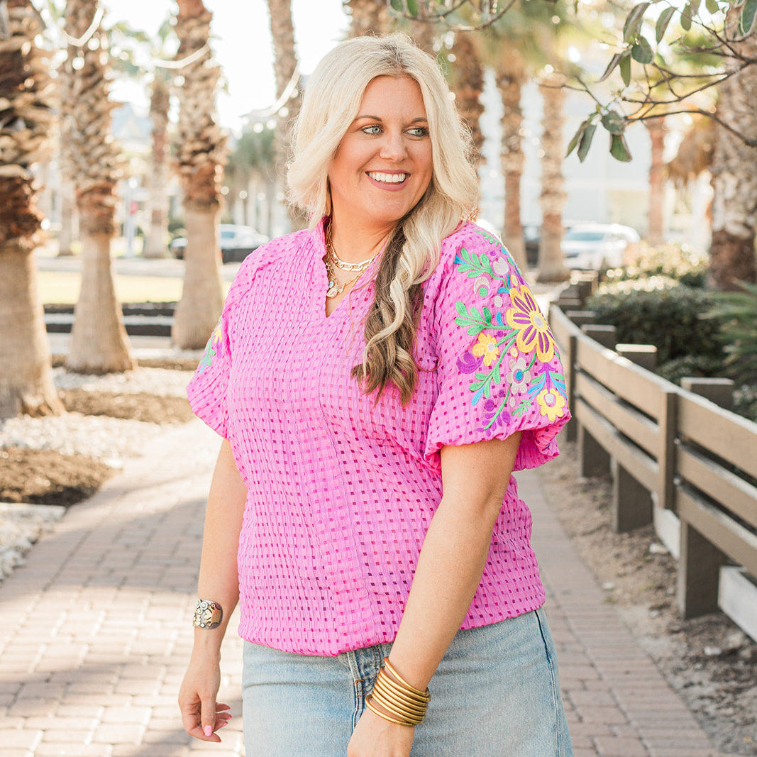 Woman with blonde hair standing in a front profile position wearing the Havanah Top in a lifestyle themed background. The focal point of this image is the top in it's entirety with emphasis on v-neckline and embroidery on sleeves. 
