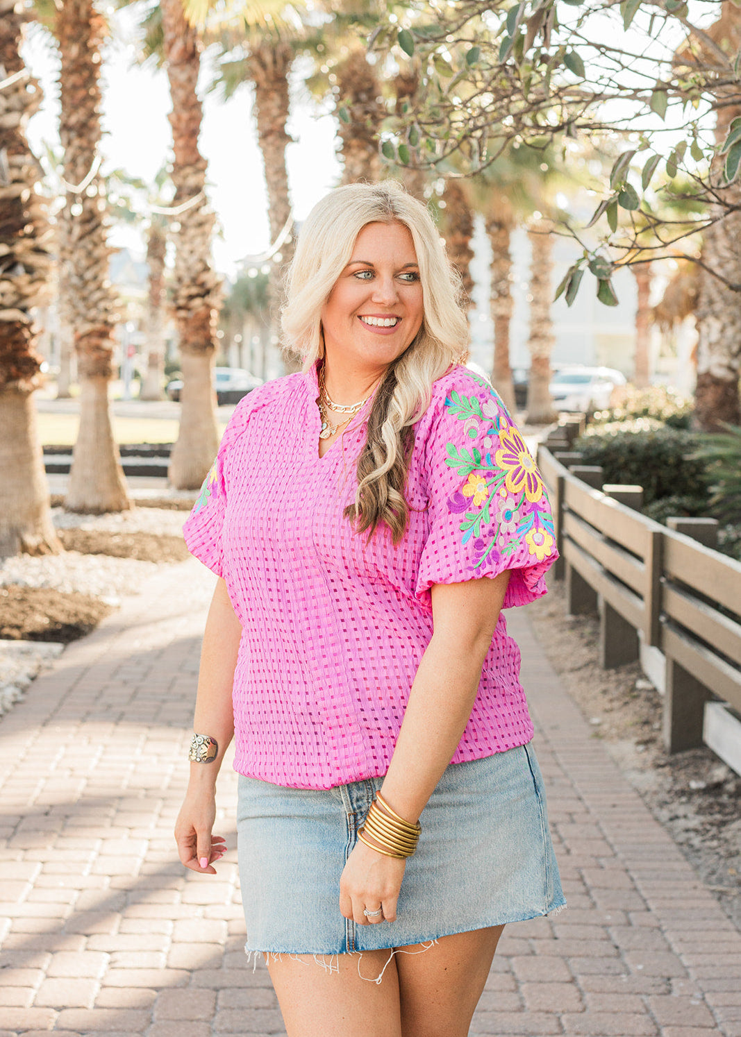 Woman with blonde hair standing in a front profile position wearing the Havanah Top in a lifestyle themed background. The focal point of this image is the top in it's entirety with emphasis on v-neckline and embroidery on sleeves. 
