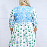 Woman with blonde hair standing in a back profile position wearing the Helen Dress against a white background. The focal point of this image is the baby blue embroidery across shoulders and gold and blue floral graphics. 