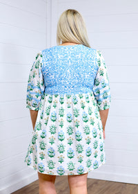 Woman with blonde hair standing in a back profile position wearing the Helen Dress against a white background. The focal point of this image is the baby blue embroidery across shoulders and gold and blue floral graphics. 