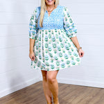Woman with blonde hair standing in a front profile position wearing the Helen Dress against a white background. The focal point of this image is the dress in its entirety (baby-blue embroidery, floral pattern, quarter sleeves, pleated cuffs). The model paired this dress with tan platform sandals. 