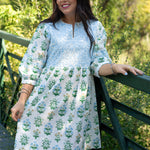 Woman with dark hair standing in a front profile position wearing the Helen Dress against in a lifestyle scene. The focal point of this image is the embroidery on chest of dress as well as v-neckline. 