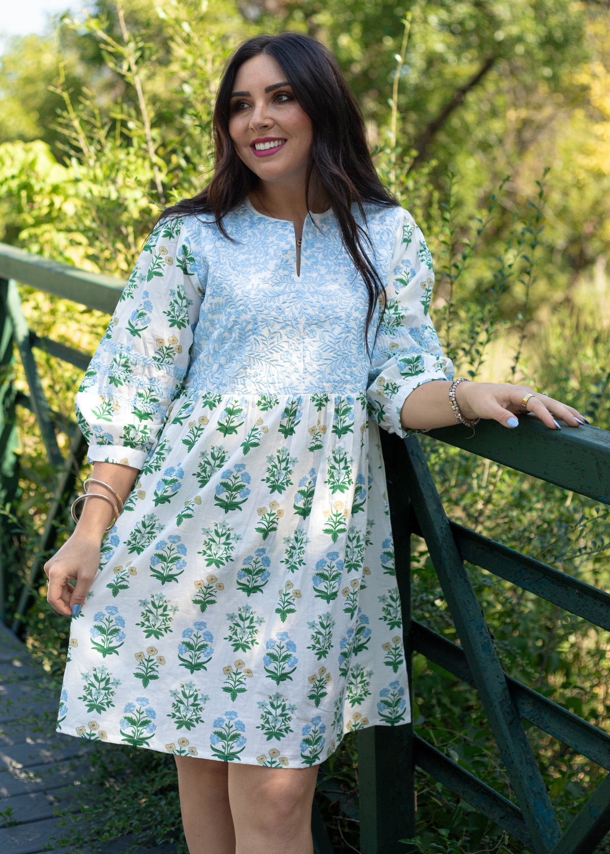 Woman with dark hair standing in a front profile position wearing the Helen Dress against in a lifestyle scene. The focal point of this image is the embroidery on chest of dress as well as v-neckline. 