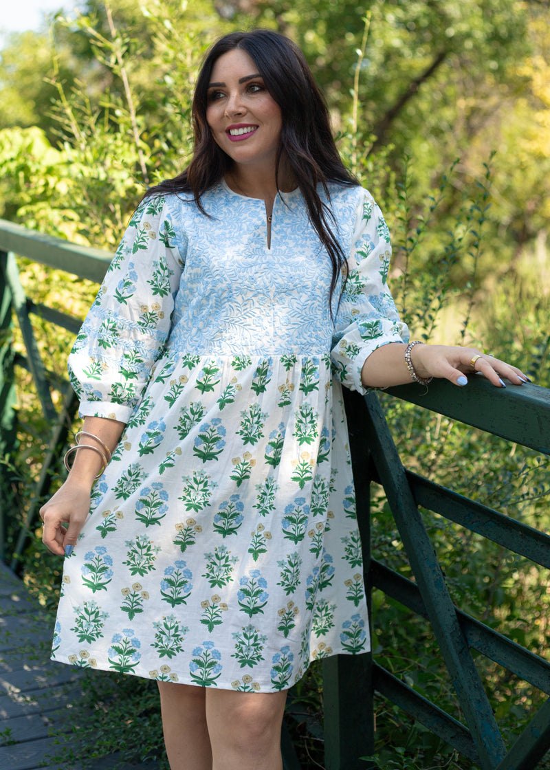 Woman with dark hair standing in a front profile position wearing the Helen Dress against in a lifestyle scene. The focal point of this image is the embroidery on chest of dress as well as v-neckline. 