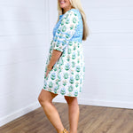 Woman with blonde hair standing in a side profile position wearing the Helen Dress against a white background. The focal point for this image is the blue and gold floral graphics on dress and baby-blue embroidery across back and shoulders. The model paired this with tan platform sandals. 