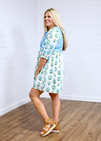 Woman with blonde hair standing in a side profile position wearing the Helen Dress against a white background. The focal point for this image is the blue and gold floral graphics on dress and baby-blue embroidery across back and shoulders. The model paired this with tan platform sandals. 