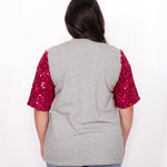 Woman with dark hair standing in a back profile position wearing the Howdy Honey Top against a white background. The focal point of this image is the pink sequin sleeves against grey fabric. The model paired this top with light-wash denim jeans. 