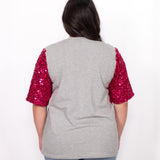 Woman with dark hair standing in a back profile position wearing the Howdy Honey Top against a white background. The focal point of this image is the pink sequin sleeves against grey fabric. The model paired this top with light-wash denim jeans. 