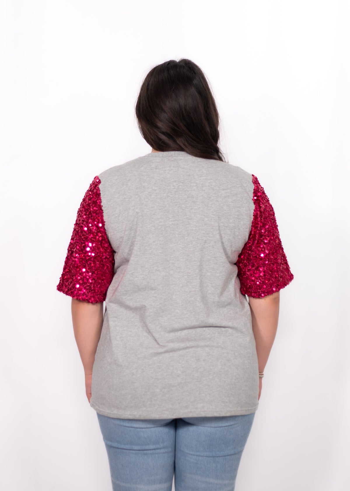 Woman with dark hair standing in a back profile position wearing the Howdy Honey Top against a white background. The focal point of this image is the pink sequin sleeves against grey fabric. The model paired this top with light-wash denim jeans. 